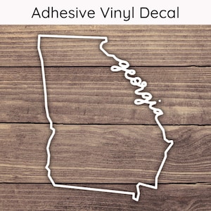 May include: White adhesive vinyl decal of the state of Georgia, with the state's name in cursive. The decal is set against a brown wood grain background. The text "Adhesive Vinyl Decal" is at the top.
