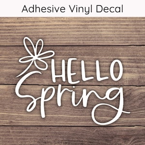 May include: White adhesive vinyl decal with the words "HELLO Spring" in a script font, with a floral accent. The decal is displayed on a wooden background. The text "Adhesive Vinyl Decal" is at the top.