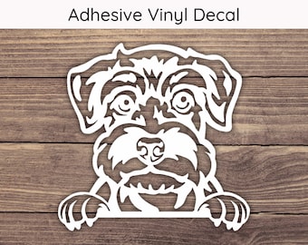 May include: White adhesive vinyl decal of a dog's face with the text "Adhesive Vinyl Decal" at the top. The dog's face is detailed with eyes, nose, and paws. The decal is on a wooden background.