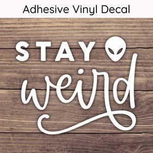May include: White vinyl decal with the text "STAY weird" and a small alien face graphic on a wood background.