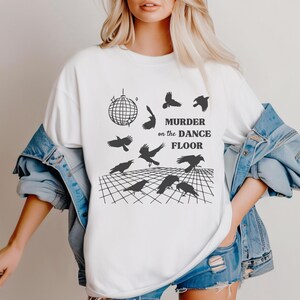 May include: White t-shirt with a black graphic design of a disco ball and crows. The text "Murder on the Dance Floor" is printed on the shirt.