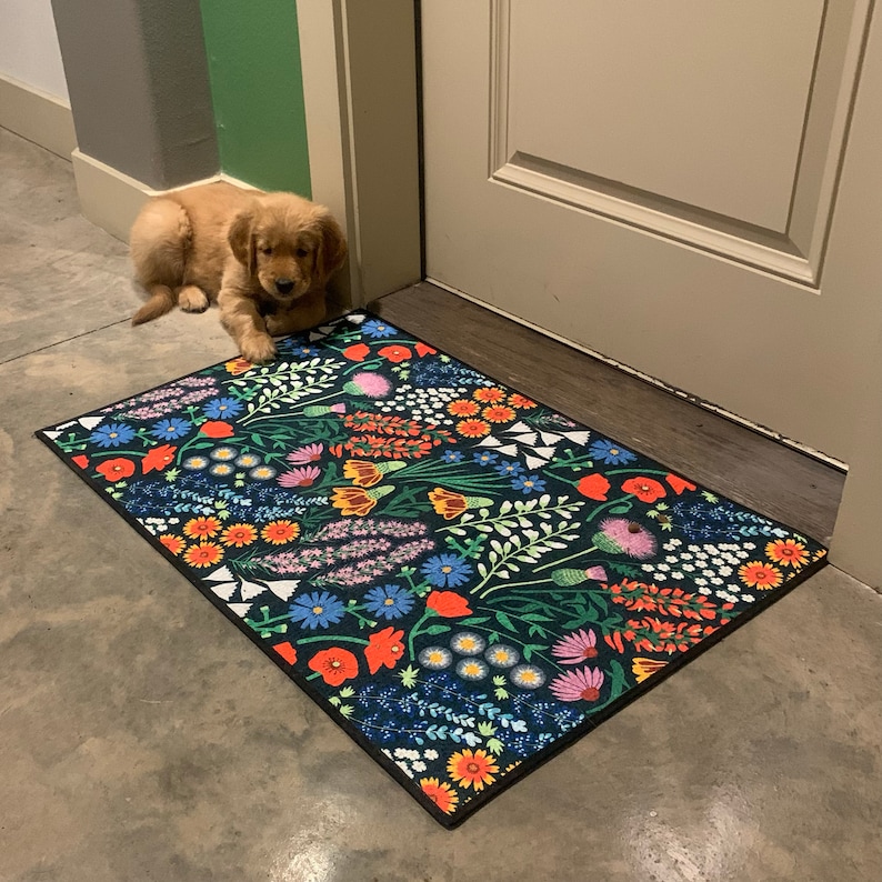 May include: A black and white floral patterned doormat with a golden retriever puppy lying on the floor next to it.