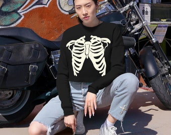 May include: Black cropped sweatshirt featuring a cream-colored rib cage design. The sweatshirt is paired with light-wash denim jeans and white sneakers. A motorcycle is visible in the background, adding a touch of urban style.