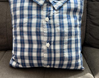 May include: A square pillow made from a blue, white, and pink plaid button-down shirt. The pillow has a collar, buttons, and a pocket. The pillow is on a gray surface.