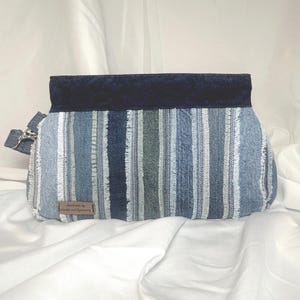 May include: A denim clutch with vertical stripes in shades of blue and white. The top flap is a solid navy blue, and a small tag reads "Handmade by Circles and Symmetry". The clutch has a silver-tone zipper and a small strap.