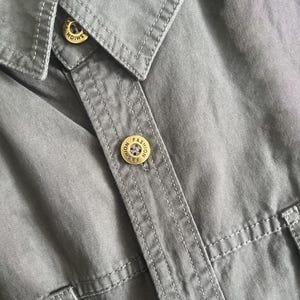 Puede incluir: Primer plano de una camisa gris con botones dorados. Los botones est&aacute;n grabados con la palabra "FASHION". La camisa tiene cuello y detalles de costura visibles.