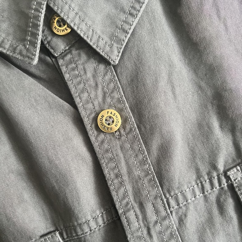 Puede incluir: Primer plano de una camisa gris con botones dorados. Los botones est&aacute;n grabados con la palabra "FASHION". La camisa tiene cuello y detalles de costura visibles.
