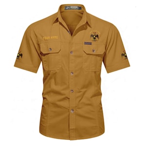 Puede incluir: Una camisa de manga corta abotonada de color beige con dos bolsillos en el pecho, cuello y emblemas de &aacute;guila bordados en el pecho y las mangas. El texto "YOUR NAME" est&aacute; bordado a la derecha y una etiqueta "Outdoor" est&aacute; en el bolsillo izquierdo.