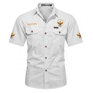 Puede incluir: Camisa blanca de manga corta con dos bolsillos en el pecho y botones. La camisa presenta texto dorado que dice "YOUR NAME" y "YOUR LIVES HERE" junto con emblemas de &aacute;guila. La camisa tiene cuello y est&aacute; etiquetada como "OUTDOOR".