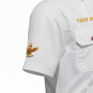 Puede incluir: Camisa blanca de manga corta con botones y dos bolsillos en el pecho. La manga izquierda presenta un emblema de &aacute;guila dorada y marr&oacute;n. El texto "YOUR NAME" est&aacute; bordado en dorado en el lado derecho.