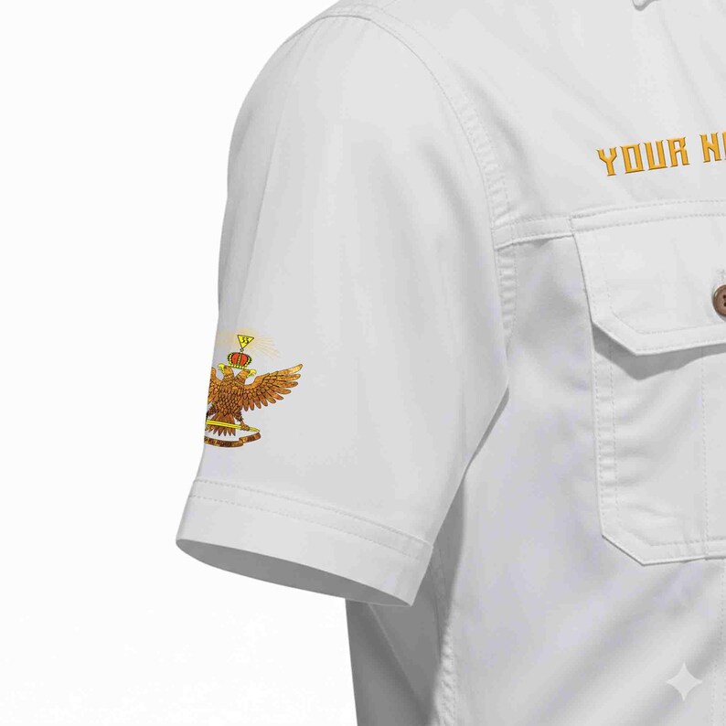 Puede incluir: Camisa blanca de manga corta con botones y dos bolsillos en el pecho. La manga izquierda presenta un emblema de &aacute;guila dorada y marr&oacute;n. El texto "YOUR NAME" est&aacute; bordado en dorado en el lado derecho.