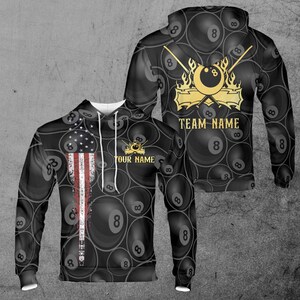 Puede incluir: Sudadera con capucha negra con un patrón repetido de bolas de billar con el número 8. La parte delantera presenta un diseño de bandera estadounidense desgastada. La parte trasera muestra un emblema dorado con tacos cruzados, una bola de billar y el texto "TEAM NAME". La parte delantera también tiene el texto "YOUR NAME".