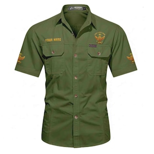 Puede incluir: Camisa verde oliva de manga corta con dos bolsillos en el pecho y cierre de botones. La camisa presenta emblemas dorados bordados y el texto "YOUR NAME" encima del bolsillo izquierdo. Tiene cuello cl&aacute;sico y etiqueta "OUTDOOR".