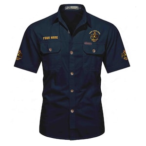 Puede incluir: Una camisa azul marino oscuro de manga corta con botones, dos bolsillos en el pecho, cuello y bordados dorados. El texto "YOUR NAME" est&aacute; impreso en el pecho derecho, mientras que un emblema de &aacute;guila bic&eacute;fala aparece en el pecho izquierdo y las mangas.