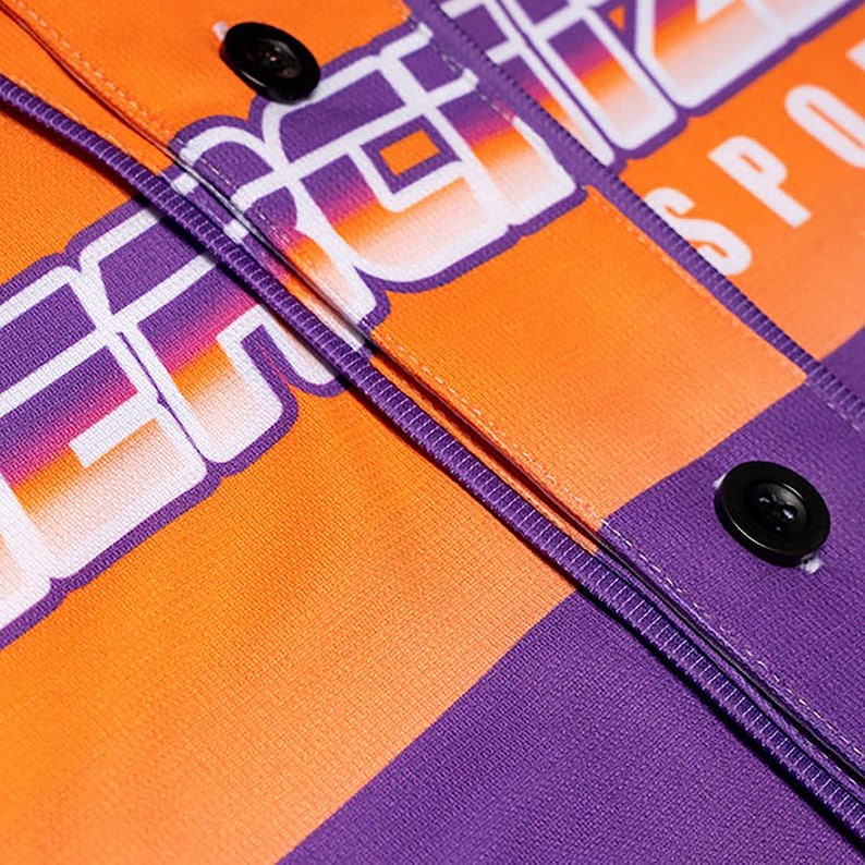 May include: Close-up of a purple and orange baseball jersey with the word "Energize" in white letters with a gradient effect. The jersey has a button closure and a purple trim.