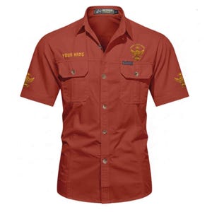 Puede incluir: Camisa roja ladrillo de manga corta con dos bolsillos en el pecho y botones. La camisa presenta detalles bordados dorados, incluyendo un emblema de &aacute;guila y los textos "YOUR NAME" y "YOUR LITTLE WING". La camisa est&aacute; etiquetada como "OUTDOOR" en la parte superior.