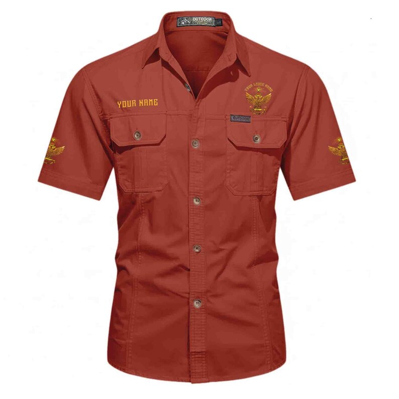 Puede incluir: Camisa roja ladrillo de manga corta con dos bolsillos en el pecho y botones. La camisa presenta detalles bordados dorados, incluyendo un emblema de &aacute;guila y los textos "YOUR NAME" y "YOUR LITTLE WING". La camisa est&aacute; etiquetada como "OUTDOOR" en la parte superior.