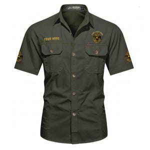Puede incluir: Una camisa de manga corta abotonada de color verde oliva con dos bolsillos en el pecho con solapa. La camisa muestra el texto "YOUR NAME" en el pecho derecho, un emblema de &aacute;guila en el pecho izquierdo y emblemas iguales en ambas mangas.