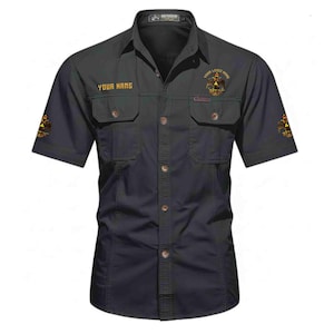 Puede incluir: Una camisa de manga corta gris oscuro con dos bolsillos en el pecho con solapas, cuello y tapeta de botones central. La camisa muestra el texto YOUR NAME y un emblema dorado de &aacute;guila bic&eacute;fala en el pecho y en ambas mangas.