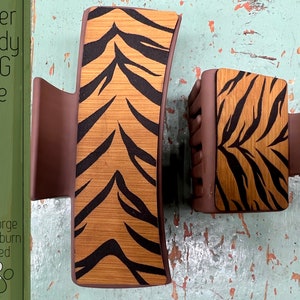 May include: Two brown hair claw clips with a tiger stripe design. The clips have a rectangular shape with a wooden tiger stripe pattern and brown plastic sides. The text "Laser Ready SVG File" and "Glowforge & Lightburn Tested" is visible.