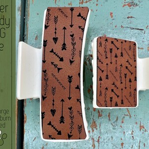 May include: Two white hair clips with brown and black arrow patterns. The larger clip is curved and the smaller clip is rectangular.