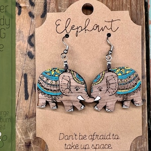 May include: A pair of wooden elephant earrings with a blue, yellow, and brown mandala design. The earrings are hanging on a brown card with the word "Elephant" written on it. The card also has the text "Don't be afraid to take up space."