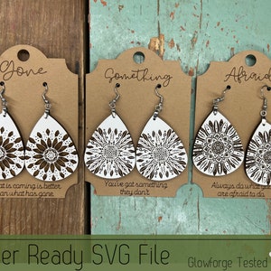 May include: Three pairs of laser-cut wooden earrings with a white mandala design. The earrings are shaped like teardrops and have silver hooks. The cards they are displayed on have the words "Gone", "Something", and "Afraid" written on them, along with inspirational quotes.