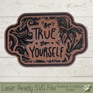 May include: A brown wooden sign with black lettering that reads "Be True to Yourself". The sign is decorated with black floral designs.