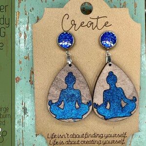 May include: A pair of wooden earrings with a blue glitter finish, featuring a silhouette of a person in a yoga pose. The earrings are hanging from silver hooks and have blue glitter embellishments. The text "Create" is visible on the background card. The text "Life isn't about finding yourself. Life is about creating yourself." is also visible on the card.