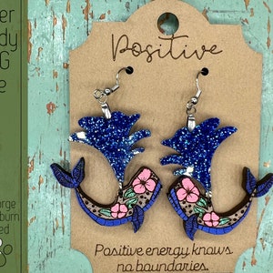 Może przedstawiać: Para niebieskich i różowych kolczyków w kształcie wieloryba z kwiatowymi akcentami. Kolczyki są wycinane laserowo i gotowe do użycia w projektach rękodzielniczych. Tekst "Positive energy knows no boundaries." jest napisany na metce pod kolczykami.