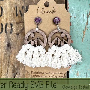 May include: A pair of wooden earrings with a mountain silhouette design and white macrame tassels. The earrings are on a brown card with the text "Climb" and "Each fresh peak ascended teaches us something new".