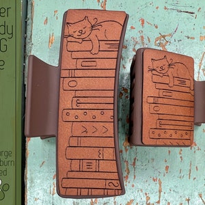 May include: Two brown hair clips with a laser-etched design of a cat sleeping on a stack of books. The text "Laser Ready SVG File" is visible in the upper left corner of the image.