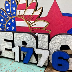 May include: Wooden sign with the words "America 1776" in white and blue paint. The sign is decorated with a red, white, and blue sunflower and stars.