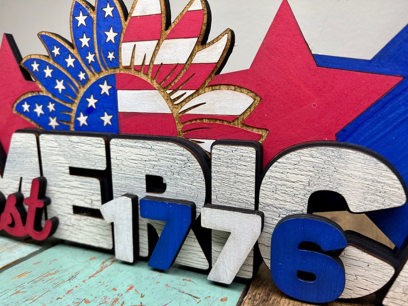May include: Wooden sign with the words "America 1776" in white and blue paint. The sign is decorated with a red, white, and blue sunflower and stars.