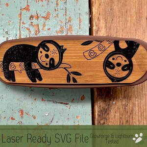 May include: A wooden rectangle with a brown border and a laser-cut design of two sloths on a branch. The sloths are black and the branch is brown. The background is a light brown wood.