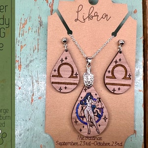 May include: A wooden jewelry set featuring the Libra zodiac sign. The set includes a necklace and earrings with a teardrop-shaped pendant and earrings. The pendant features a woman holding a scale, while the earrings feature the Libra symbol. The text "The Balance September 23rd-October 23rd" is printed below the pendant.