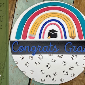 Peut inclure: Panneau de graduation rond en bois avec un arc-en-ciel coloré et l'inscription bleue "Congrats Grad". L'arc-en-ciel présente des arcs turquoise, roses, jaunes et bleus, avec une toque et des crayons. Le bas est blanc avec des icônes de chapeaux de graduation.