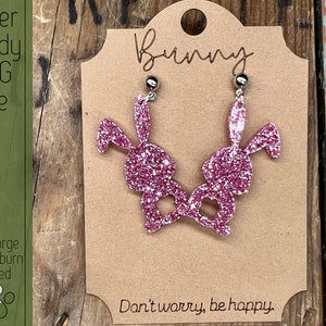May include: A pair of pink glitter bunny earrings with silver ear wires. The earrings are displayed on a brown card with the text "Bunny" and "Don't worry, be happy."