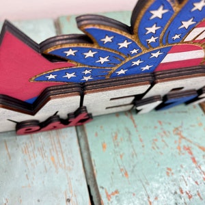 May include: Wooden sign with the words "Happy 4th" in red, white, and blue. The sign features a red, white, and blue firework with stars.