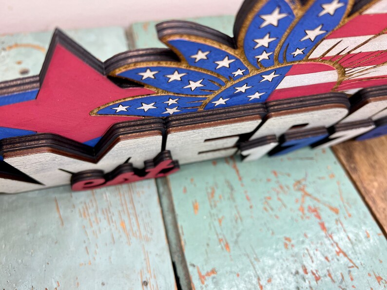 May include: Wooden sign with the words "Happy 4th" in red, white, and blue. The sign features a red, white, and blue firework with stars.