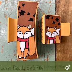 May include: Two orange hair claw clips with a fox design. The fox is wearing a red scarf and is set against a brown background with black leaf accents. The text "Laser Ready SVG File" is at the bottom.