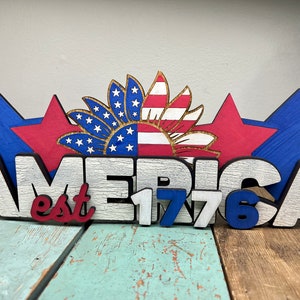 May include: Wooden sign with the word "AMERICA" in white letters with black outlines, and the text "est 1776" in red, white, and blue letters. The sign is decorated with three red and blue stars and a sunflower with red and white stripes.