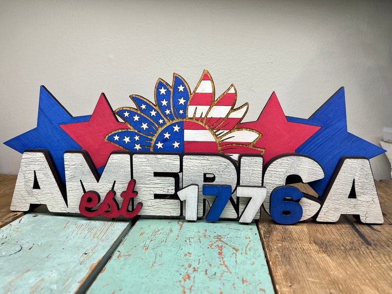 May include: Wooden sign with the word "AMERICA" in white letters with black outlines, and the text "est 1776" in red, white, and blue letters. The sign is decorated with three red and blue stars and a sunflower with red and white stripes.
