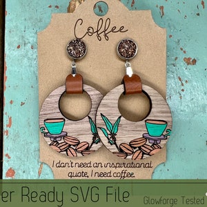 May include: A pair of wooden earrings with a coffee cup and coffee beans design. The earrings are brown with a light wood grain and have a silver hook. The earrings are on a brown card with the text "Coffee" and "I don't need an inspirational quote, I need coffee."