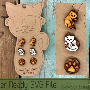 Pode incluir: Um conjunto de brincos e um alfinete com tema de gato, exibidos em um suporte de papelão em forma de gato. Os brincos apresentam desenhos de gatos laranja e brancos, juntamente com pegadas. O texto "One Cat short of Crazy" é visível.