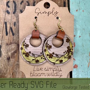 May include: Wooden hoop earrings with gold-colored floral accents and leather accents. The earrings are laser-cut with a floral design and hang from gold-colored hooks. The earrings are attached to a brown card with the text "Simply" and "Live simply, bloom wildly."