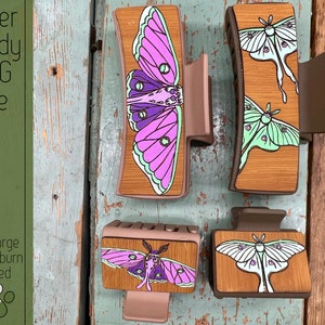 Può includere: Quattro mollette per capelli in legno con disegni di falene tagliati al laser. Le falene sono in tonalità di rosa, viola, verde e bianco. Il testo "Laser Ready SVG File" e "Glowforge & Lightburn Tested" è visibile sul lato sinistro dell'immagine.