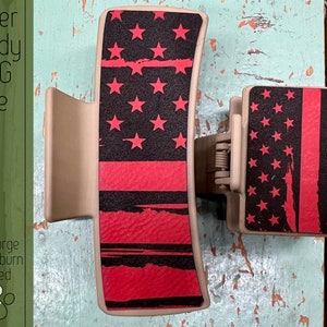 May include: A red, white, and blue American flag design on a hair clip. The clip is made of a beige plastic material. The design is a distressed look with a red stripe and white stars on a black background.