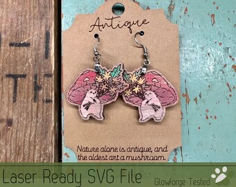 May include: A pair of laser-cut wooden earrings featuring a mushroom with a small animal holding a bouquet of flowers. The earrings are pink, green, and yellow. The text "Antique" and "Nature alone is antique, and the oldest art a mushroom." is printed on the card.