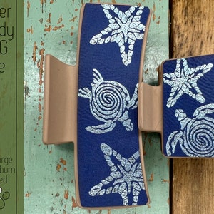May include: A blue hair clip with a white design of sea turtles and starfish. The clip is made of plastic and has a metal spring.